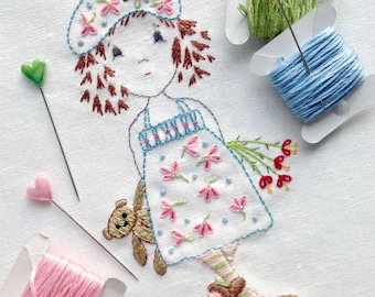 May include: Embroidered image of a girl with brown hair wearing a blue dress and a blue and white hat. She is holding a brown teddy bear and a bouquet of red flowers. The image is surrounded by spools of thread and pins.