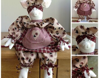 May include: A handmade pig doll wearing a brown and white dress with a red and white gingham apron. The doll has a white face with black eyes and a pink nose.