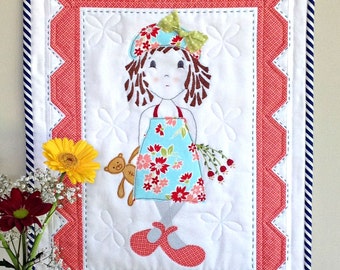 May include: A white quilted wall hanging with a red and white border. The center features an embroidered image of a girl with brown hair wearing a blue dress, a red hat, and red shoes. She is holding a teddy bear and a bouquet of flowers.