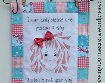 May include: A white fabric wall hanging with a patchwork border of pink and blue gingham and floral print fabric. The center of the hanging features a girl with pink hair and a red bow, and the text "I can only please one person a day. Today is not your day."