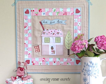 May include: A handmade quilted wall hanging with a pink and white house, a tree, and a banner that reads "love lives here". The background is a beige fabric with a pink and white checkered border. The hanging is titled "sewing room secrets".