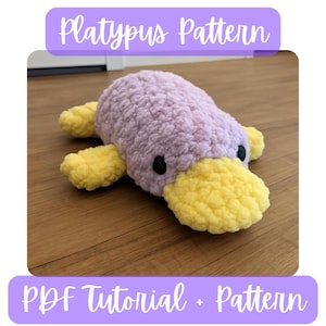 Puede incluir: Un juguete de ornitorrinco tejido a ganchillo en tonos lavanda y amarillo. El animal de peluche tiene un cuerpo texturizado, un pico amarillo y pequeños ojos negros. La imagen incluye el texto "Platypus Pattern" y "PDF Tutorial + Pattern".