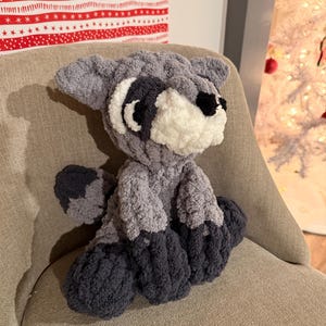 May include: A handmade plush raccoon toy in shades of gray and black, with a white muzzle and black eyes. The stuffed animal is sitting on a beige chair. The raccoon has a fluffy texture and a cute, friendly appearance.