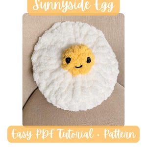 May include: A white crocheted plush egg with a yellow center featuring a stitched black face. The text Sunnyside Egg appears at the top, and Easy PDF Tutorial + Pattern is at the bottom. The egg has a soft, textured, fuzzy yarn appearance.