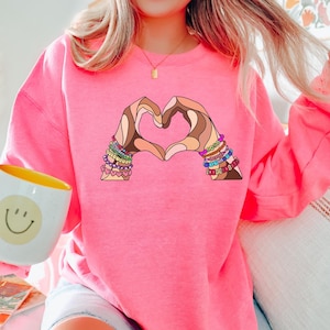 May include: Pink sweatshirt with a graphic of two hands forming a heart shape. The hands are adorned with colorful bracelets.