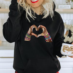 Op de afbeelding: Zwart hoodie met een afbeelding van twee handen die een hartvorm vormen. De handen zijn veelkleurig en hebben armbanden.