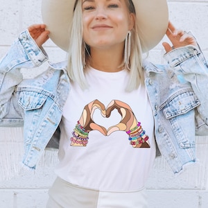 May include: White t-shirt featuring a heart shape formed by two hands with various skin tones. The hands are adorned with colorful bracelets. The person is wearing a light blue denim jacket and a beige hat.