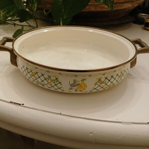 Może przedstawiać: Owalne naczynie do pieczenia z emalii z białym wnętrzem i ozdobnym obramowaniem z zielono-żółtym wzorem kratki z ilustracjami owoców. Naczynie ma uchwyty i wykończenia w kolorze brązu.