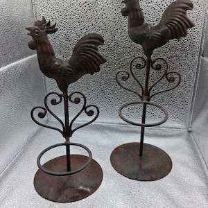 Puede incluir: Dos figuras de gallo de metal rústico. Cada gallo está posado sobre un soporte decorativo con una base circular y un diseño en forma de corazón. Los gallos y los soportes tienen un acabado marrón desgastado.
