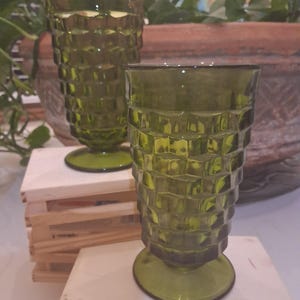 May include: Two olive green drinking glasses with a textured, geometric pattern. The glasses have a pedestal base and a tall, cylindrical shape. The glasses are made of glass and are likely vintage. The glasses are displayed on a wooden surface.