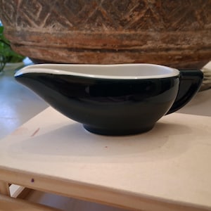 May include: A dark teal ceramic gravy boat with a white interior and a handle. The gravy boat is on a light-coloured wooden surface. A large, textured, brown decorative pot is in the background.