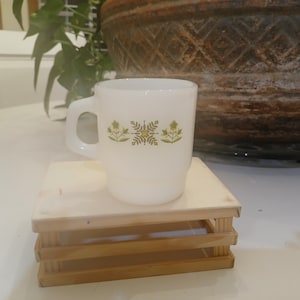 Könnte beinhalten: Eine weiße Tasse mit Henkel und grünem Blumendesign, die auf einer kleinen Holzkiste steht. Die Tasse steht vor einem verschwommenen Hintergrund mit einem großen dekorativen Topf und Grünzeug.
