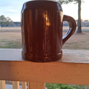 Może przedstawiać: Brązowy ceramiczny kufel do piwa z uchwytem. Kubek jest cylindryczny z lekko rozszerzoną podstawą i ciemnym, błyszczącym wykończeniem. Kubek stoi na białej drewnianej balustradzie.