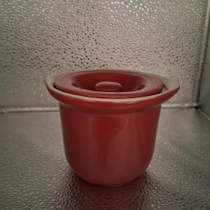 May include: A small, round, red ceramic pot with a matching lid. The pot has a slightly flared rim and a smooth, glossy finish. The lid has a recessed center and a small handle. The pot is likely used for cooking or serving.