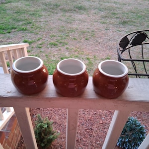Könnte beinhalten: Drei kleine, runde, braune Keramiktöpfe mit weißen Innenräumen und Rändern sind auf einem Holzgeländer angeordnet. Die Töpfe haben eine glänzende Oberfläche. Der Hintergrund umfasst eine grasbewachsene Fläche und einen dekorativen Metallkorb.