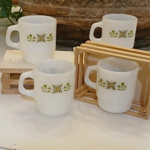 Könnte beinhalten: Vier weiße Keramikbecher mit Henkel und einem floralen Design in Grün und Gelb. Die Becher sind auf einer kleinen Holzpalette und in einer Holzkiste gestapelt.