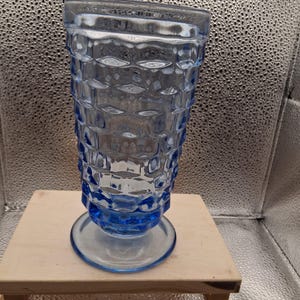 May include: A vintage blue glass vase with a textured, geometric pattern. The vase has a flared top, a wide base, and a pedestal foot. The glass is a light, transparent blue, and the vase is sitting on a small wooden crate.