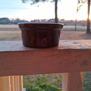 Op de afbeelding: Een kleine, donkerbruine keramische kom met een glanzende afwerking. De kom is rond met een licht uitlopende rand. Hij staat op een wit geschilderd houten oppervlak, met een vage buitenachtergrond van bomen en een zonsondergang.