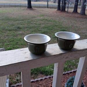 Może przedstawiać: Dwie oliwkowozielone miski emaliowane z białym wnętrzem i czarnymi obwódkami stoją na zwietrzałej białej balustradzie. Miski mają prosty, klasyczny design, idealny do serwowania lub ekspozycji. Miski znajdują się na zewnątrz, na trawiastym tle.