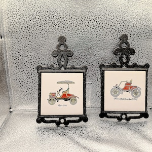 Può includere: Due sottopentola con auto d'epoca. Ognuno ha una cornice in metallo nero con elementi decorativi. Le piastrelle mostrano illustrazioni colorate di auto d'epoca, con testo che indica il modello e l'anno.