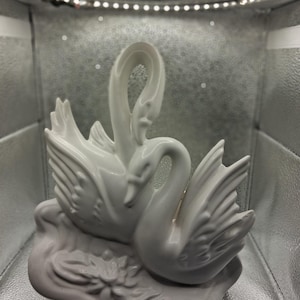May include: A white ceramic figurine of two swans, intertwined in a graceful pose. The swans have detailed feather textures and are perched on a base with floral accents. The sculpture is a decorative item.