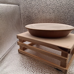 May include: A brown ceramic oval serving dish with a cream interior rests on a small, light-coloured wooden stand. The dish has a simple, classic design. The stand is made of unfinished wood.