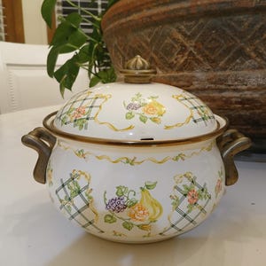 Może przedstawiać: Biały garnek emaliowany z pokrywką, z kwiatowym i owocowym wzorem. Garnek ma mosiężne uchwyty i wykończenia. Pokrywka zwieńczona jest mosiężnym guzikiem. Garnek stoi na białej powierzchni.