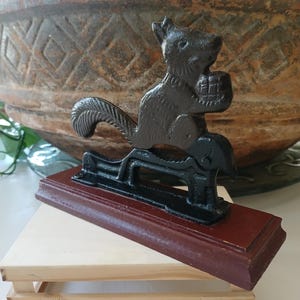 May include: A cast iron squirrel nutcracker, painted black, holding a nut. The squirrel sits atop a lever mechanism mounted on a rectangular, dark red wooden base. The base is supported by a small, light-colored wooden stand.