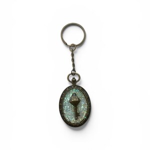 Può includere: Un portachiavi di forma ovale con una cornice in metallo color bronzo antico. Il centro contiene un inserto in resina chiara riempito di glitter iridescenti. Un piccolo ciondolo a forma di chiave color bronzo è incastonato al centro.