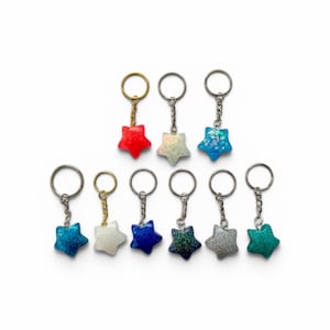 May include: A collection of nine star-shaped keychains, each featuring a unique glittery or solid finish in colors including red, white, blue, silver, and teal. Each star is attached to a metal split ring by a short silver or gold-toned chain.