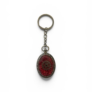 May include: An oval-shaped keyring featuring a bronze-toned metal frame with an ornate border. The centre contains a deep red, glittery resin inlay embedded with a single, small, dark bronze-coloured gear. It includes a matching metal chain and ring.