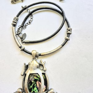 May include: A silver-toned frog pendant necklace with matching frog earrings. The frog pendant and earrings have a colorful iridescent inlay. The necklace has a black cord and silver accents.