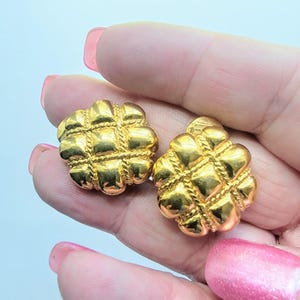 May include: A pair of gold-colored, textured stud earrings. Each earring features a quilted design with rope-like detailing. The earrings are round and have a shiny finish. The earrings are held in a hand with pink nail polish.