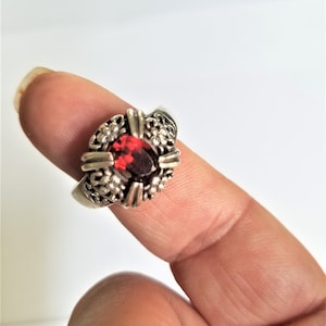 May include: Silver ring with a large, oval, deep red gemstone. The ring features intricate filigree and floral detailing around the gemstone and on the band. Gold-colored accents are visible on the ring.