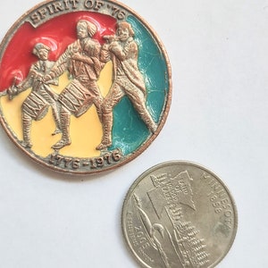 Può includere: Una moneta rotonda di colore rame con un design in smalto rosso, giallo e verde. Tre figure sono raffigurate al centro, che suonano strumenti musicali. La moneta riporta la scritta "Spirit of '76" e "1776-1976".
