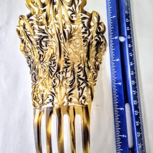 May include: A decorative hair comb with a tortoiseshell pattern featuring an intricate openwork filigree design of scrolling leaves and a bow at the top. A blue ruler is placed alongside the comb, showing a total length of approximately 8 inches.
