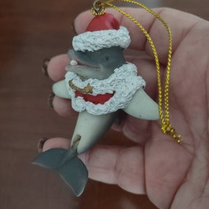 May include: A gray and white dolphin wearing a red Santa hat with white fur trim and a white fur coat with a red trim. The dolphin is holding a white pearl in its mouth. The ornament has a gold string.