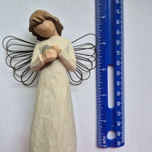 May include: A carved wooden angel figurine with a cream-colored dress and brown hair. The angel holds a small gray object and has wire wings. A blue ruler is next to the angel, showing measurements in inches.