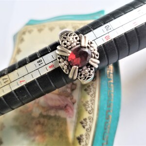 May include: Silver ring with a large, round, deep red gemstone at the center. The ring has a floral design around the gemstone. The ring is on a black ring sizer with measurements in inches.