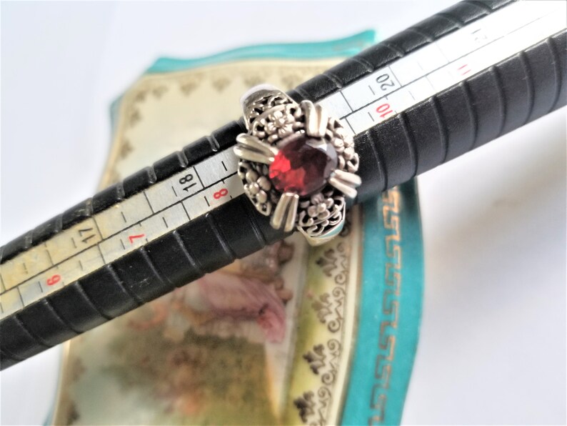 May include: Silver ring with a large, round, deep red gemstone at the center. The ring has a floral design around the gemstone. The ring is on a black ring sizer with measurements in inches.