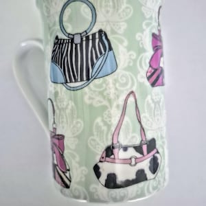 Puede incluir: Una taza de cerámica con fondo verde claro y un patrón de damasco blanco. La taza está decorada con varias ilustraciones de bolsos, incluyendo un bolso de rayas blancas y negras, un bolso rosa y negro, y un bolso con estampado de vaca blanco y negro.