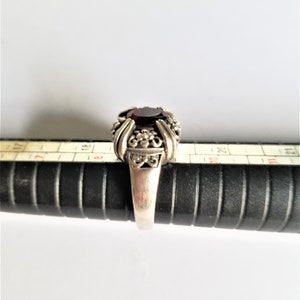May include: A silver ring with a large, oval, dark red gemstone. The ring has an ornate design with floral and scroll details around the gemstone. The ring is on a black ring sizer.