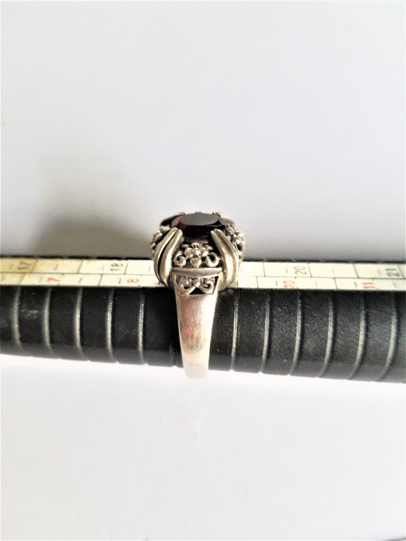 May include: A silver ring with a large, oval, dark red gemstone. The ring has an ornate design with floral and scroll details around the gemstone. The ring is on a black ring sizer.