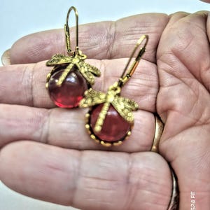 Puede incluir: Un par de pendientes de libélula dorados. Cada pendiente presenta una cuenta redonda de vidrio rojo, engastada en un marco decorativo dorado. El diseño de la libélula es visible encima de la cuenta, añadiendo un toque inspirado en la naturaleza.