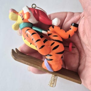 May include: A Tigger Christmas ornament. The orange and black striped Tigger figure is wearing a red Santa hat and skiing on a wooden ski. The ornament has a gold-colored metal spring and a loop for hanging.