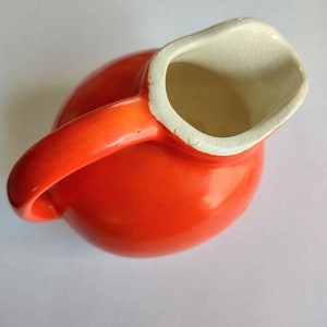 Könnte beinhalten: Eine leuchtend orangefarbene Keramikkrug mit rundem Körper und gebogenem Griff. Der Innenraum und der Rand sind cremefarben. Der Krug ist zum Ausgießen von Flüssigkeiten bestimmt und hat eine klassische, funktionale Form.