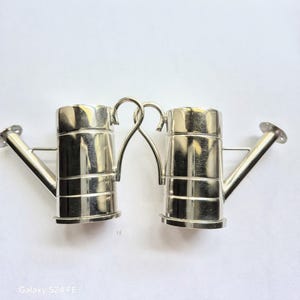 May include: Two silver-colored miniature watering cans. Each can has a cylindrical body with a spout and a handle. The handles are connected, forming a decorative arch. The watering cans are designed with a classic, vintage aesthetic.