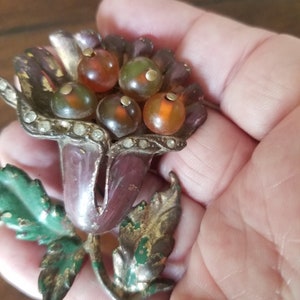 Puede incluir: Un broche vintage en forma de flor con un grupo de cuentas de vidrio ámbar y verdes en el centro. La flor está hecha de metal con un acabado morado y dorado y tiene un tallo de hoja verde.