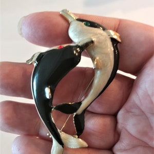 May include: A black and white enamel dolphin brooch with red and green rhinestones for eyes. The dolphins are intertwined in a playful pose.