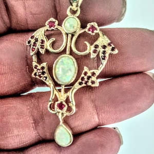 May include: A silver-toned pendant featuring a central oval opal, a smaller round opal at the top, and a teardrop-shaped opal at the bottom. The metal frame is decorated with small, deep red gemstones arranged in floral-inspired patterns.